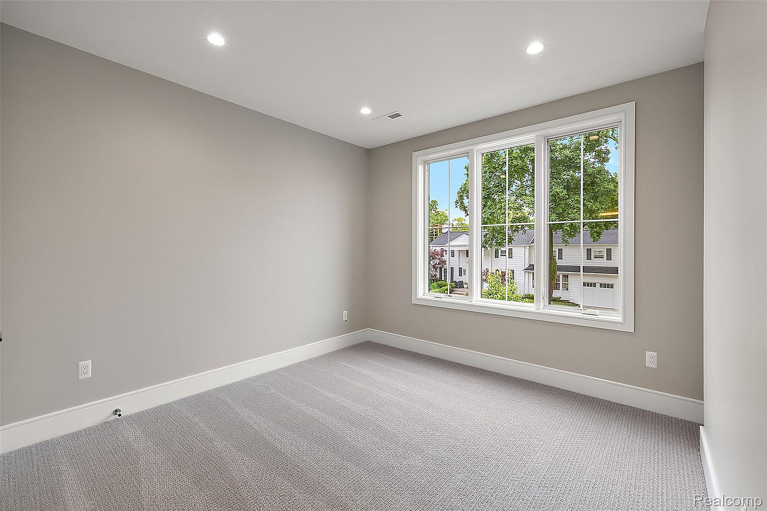The image showcases a well-lit, neutral-toned bedroom, likely a guest or secondary bedroom. The room features light gray walls, white trim, and a light gray carpet. A large window provides ample natural light and a view of the neighborhood, enhancing the room's appeal and spacious feel.