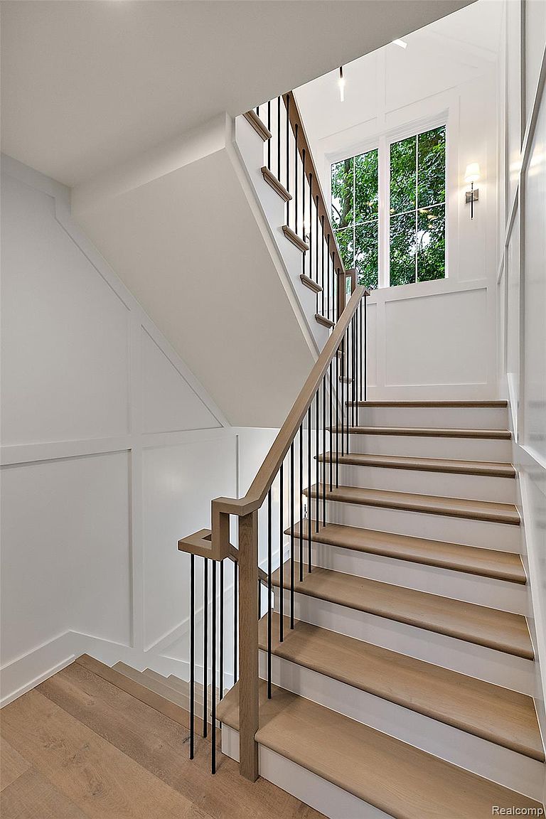 This interior shot showcases a well-lit staircase with light wood treads and white risers, complemented by a wooden handrail with black metal balusters. The walls are painted white with decorative paneling, and a large window at the top of the stairs provides natural light, enhancing the bright and airy feel of the space. The perspective is from the bottom of the stairs looking up.