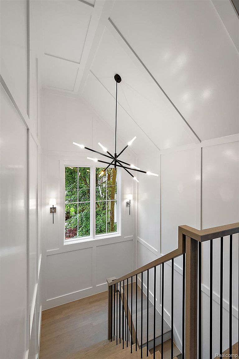 This is an interior shot of a bright hallway and staircase area. The walls are painted white and feature wainscoting details, complemented by a modern chandelier hanging from the vaulted ceiling. A window provides natural light and a view of the outdoors, while the staircase features a wooden handrail with black metal balusters, creating a clean and elegant transition between floors.