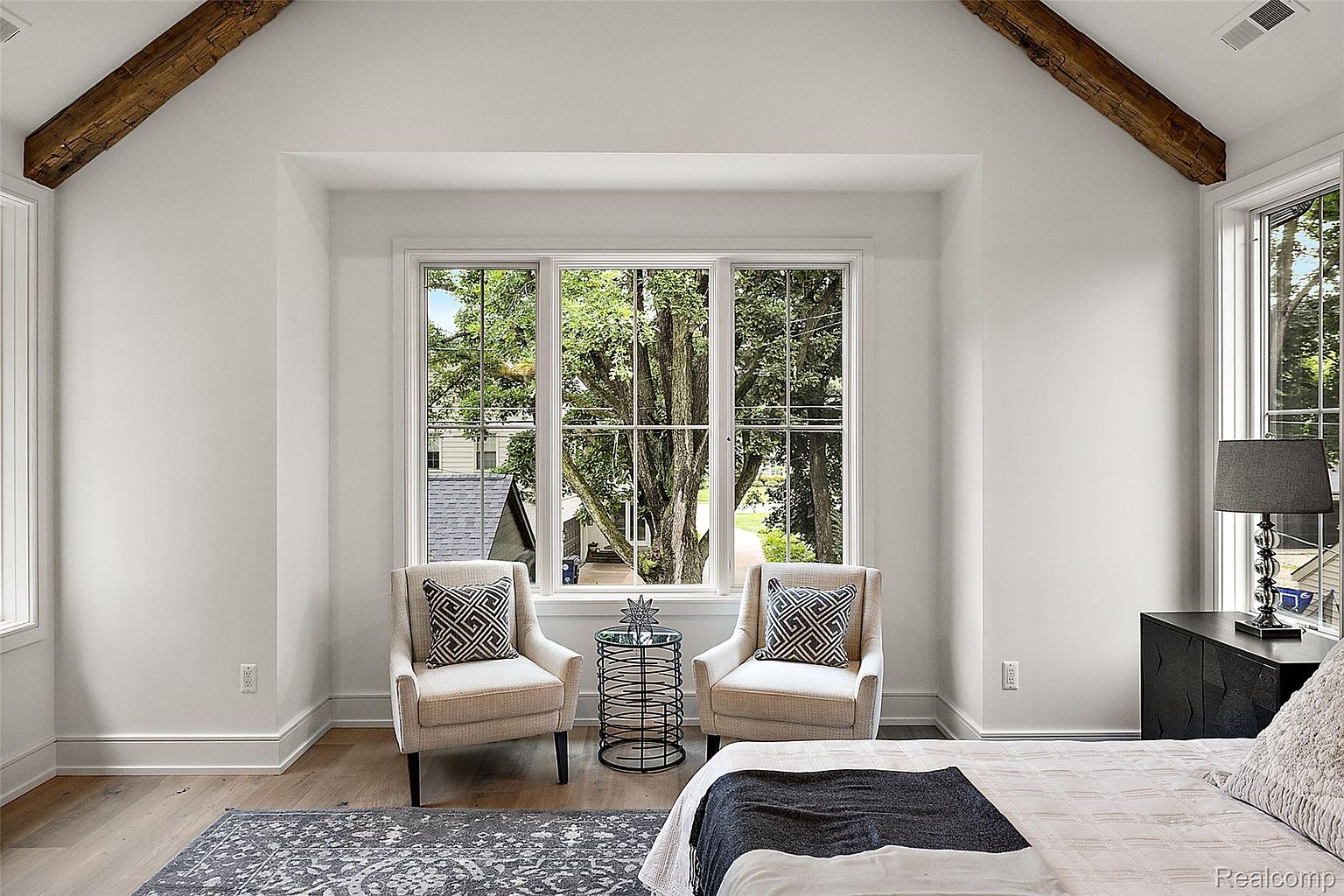This is a bright and airy primary bedroom featuring a large window with a view of mature trees. The room is decorated in neutral tones with two armchairs flanking a small table in front of the window, creating a cozy seating area. Exposed wooden beams add a touch of rustic charm to the otherwise modern space.