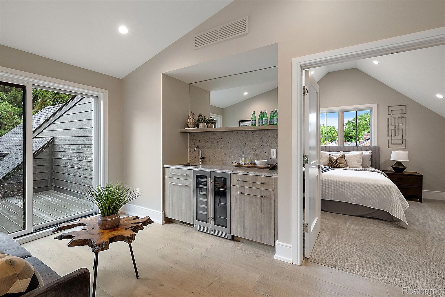 This interior shot showcases a guest bedroom adjacent to a small bar area. The bedroom features a neutral color palette, a comfortable bed with layered bedding, and a window providing natural light. The bar area includes a sink, beverage cooler, and mirrored backsplash, adding a touch of luxury and convenience to the space.