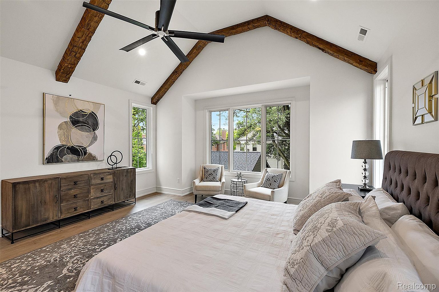 This is a primary bedroom featuring a vaulted ceiling with exposed wooden beams and a neutral color palette. The room is well-lit with natural light from multiple windows, and furnished with a large bed, a wooden dresser with artwork above, and two armchairs creating a cozy seating area. The overall impression is one of comfort and sophistication.