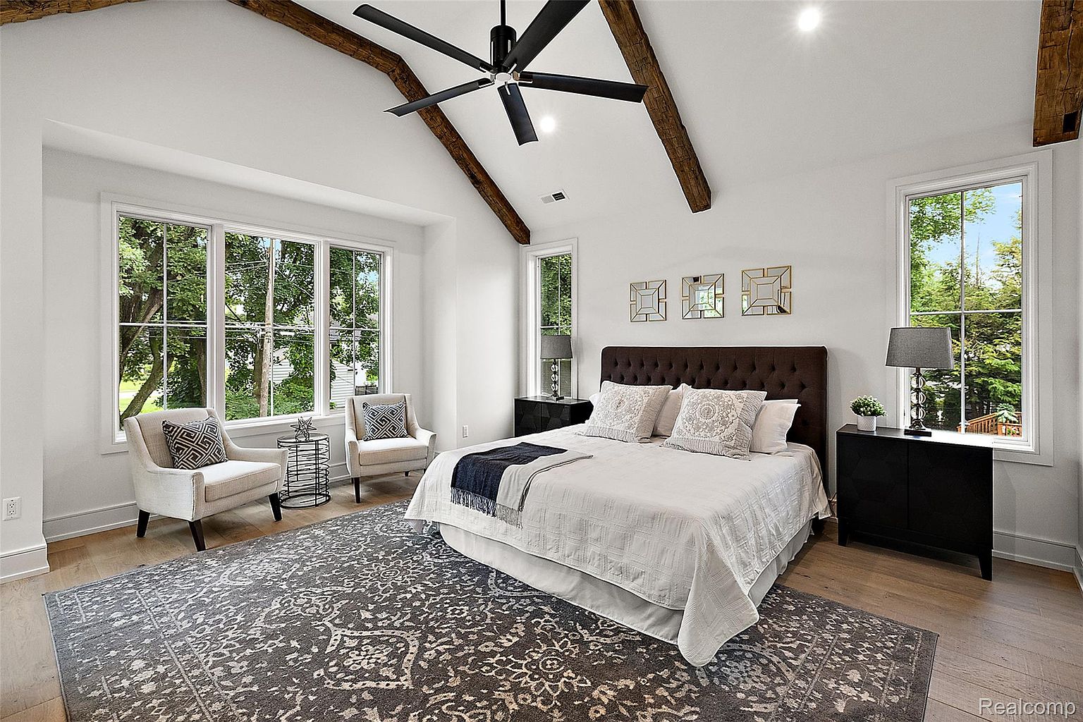 This is a primary bedroom featuring a king-size bed with a tufted headboard, flanked by matching nightstands and lamps. Large windows provide natural light and views of the outdoors, while exposed wooden beams add a rustic touch to the vaulted ceiling. The room is decorated in neutral tones with a patterned rug, creating a serene and inviting atmosphere.