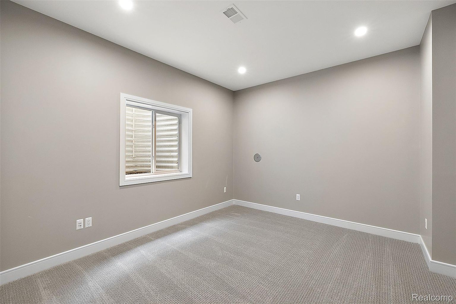 This is a well-lit, finished basement room with neutral gray walls and carpet. A window provides natural light, and recessed lighting illuminates the space. The room is clean and appears to be newly renovated, offering a blank canvas for various uses.