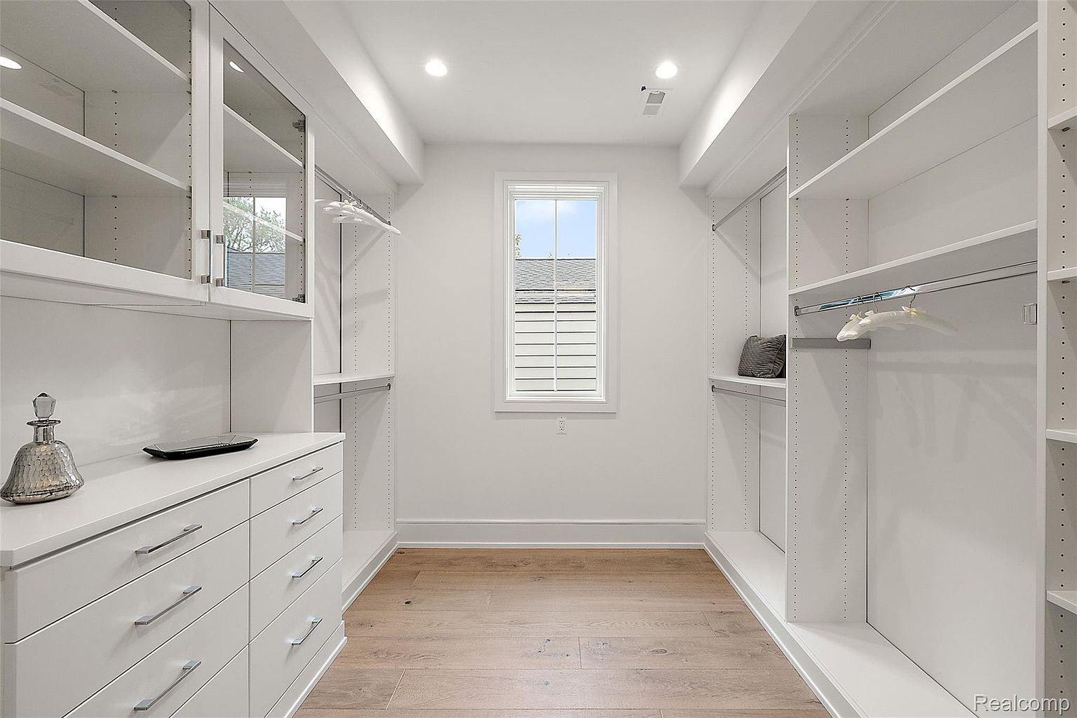 This is a well-organized walk-in closet featuring custom white shelving and drawers. The closet is brightly lit with recessed lighting and natural light from a central window. The hardwood flooring adds a touch of warmth to the space, creating a clean and luxurious feel.