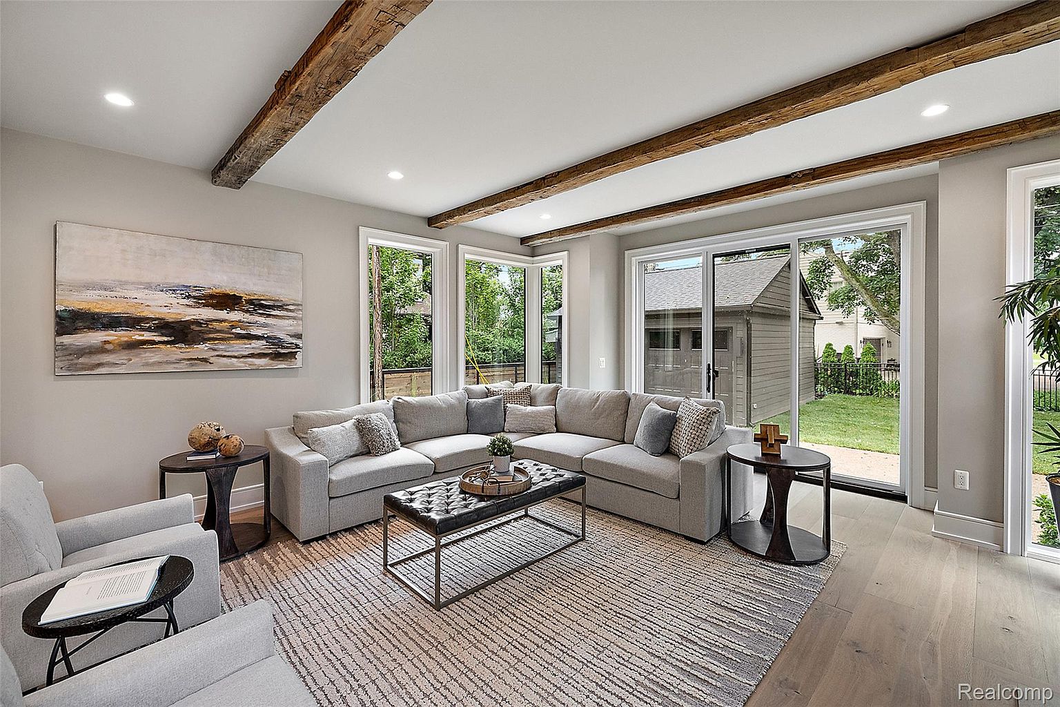 This is an interior shot of a living room featuring a large sectional sofa, a patterned area rug, and wooden ceiling beams. Natural light floods the space through large windows and sliding glass doors, offering views of the exterior. The room is decorated with neutral tones and modern furniture, creating a comfortable and inviting atmosphere.