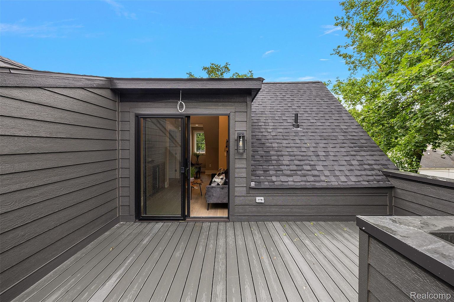 This image showcases a modern deck or balcony area with gray wood planks and dark gray siding. A sliding glass door provides access to the interior, and a section of the roof is visible, adding architectural interest. The space appears clean and well-maintained, offering a private outdoor retreat.
