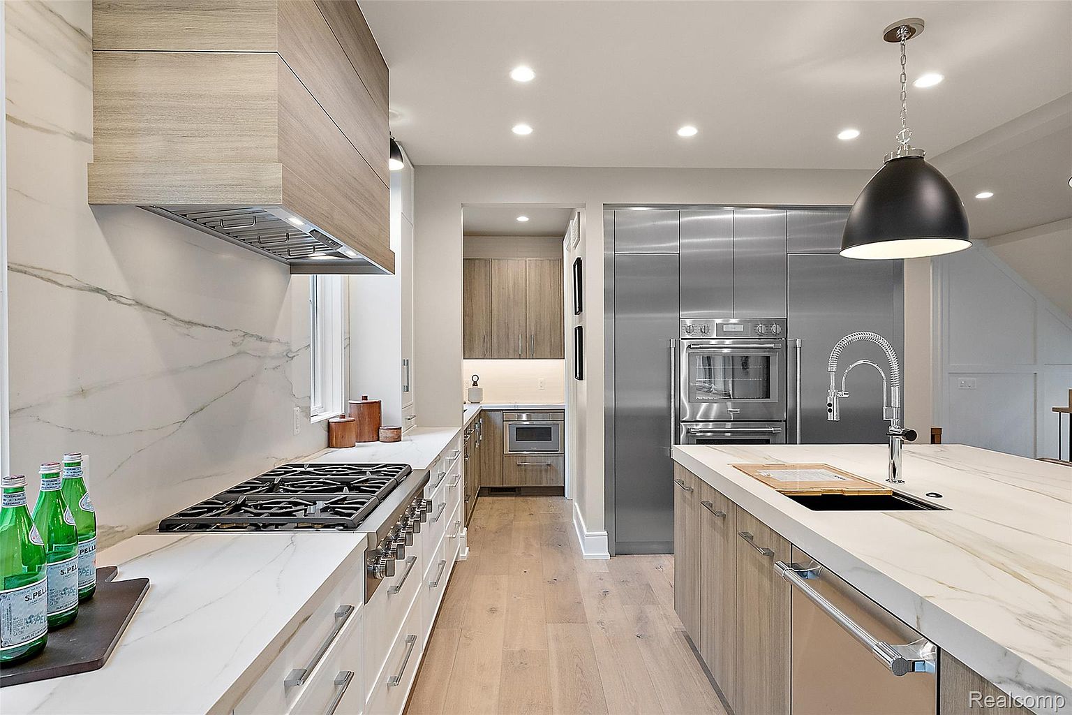 This is a modern kitchen featuring a large island with a marble countertop and a stainless steel sink. The kitchen is equipped with stainless steel appliances, including a built-in oven and refrigerator. The cabinetry is a mix of light wood and white, creating a bright and airy feel.
