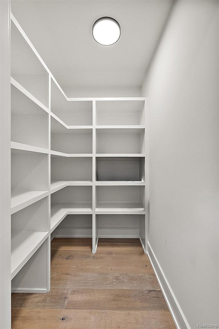 This is a well-organized pantry featuring custom white shelving and light wood flooring. The shelves are empty, providing ample storage space. A recessed light fixture illuminates the space, creating a clean and bright look.