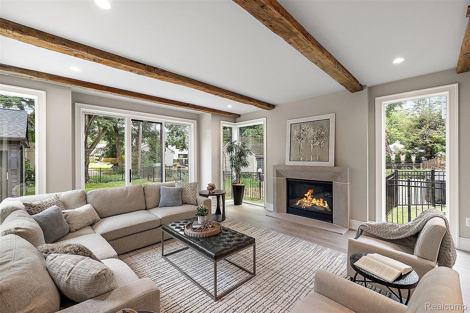 This is an interior shot of a living room featuring a large sectional sofa, a tufted ottoman coffee table, and a fireplace with a stone surround. The room has wooden ceiling beams and large windows that provide natural light and views of the exterior. The overall style is modern and inviting, creating a comfortable and stylish living space.