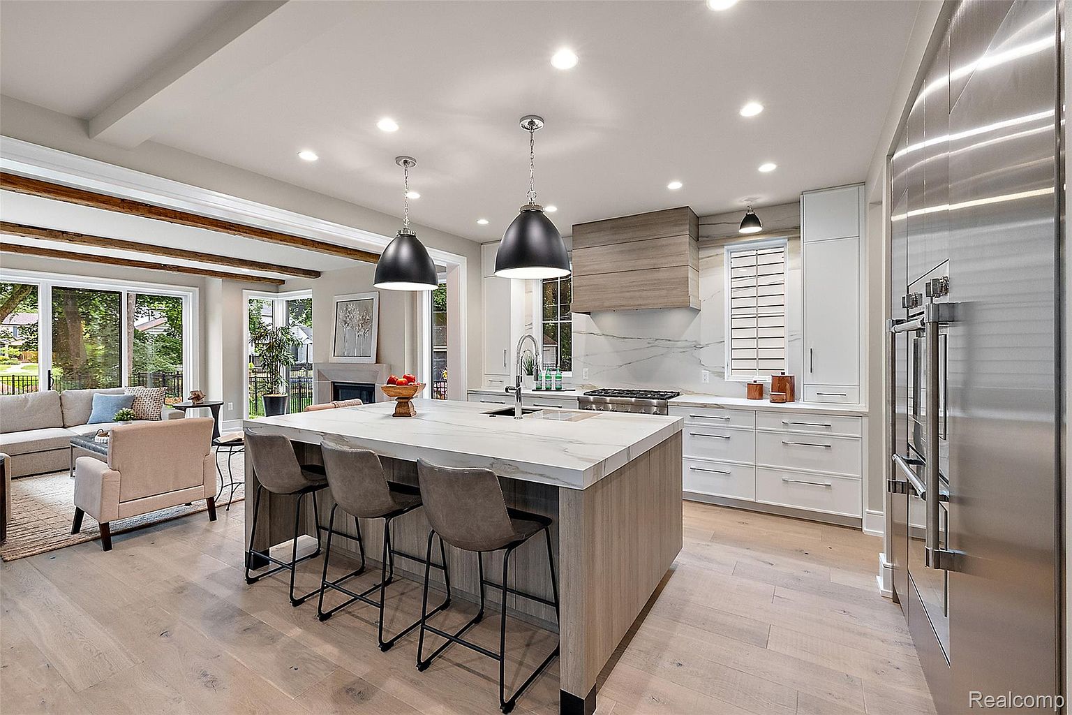 This is a bright and modern kitchen featuring a large island with seating, pendant lighting, and stainless steel appliances. The kitchen has white cabinetry and a light wood floor, creating a clean and airy feel. The open-concept design connects the kitchen to a living area with large windows, enhancing the sense of space and light.