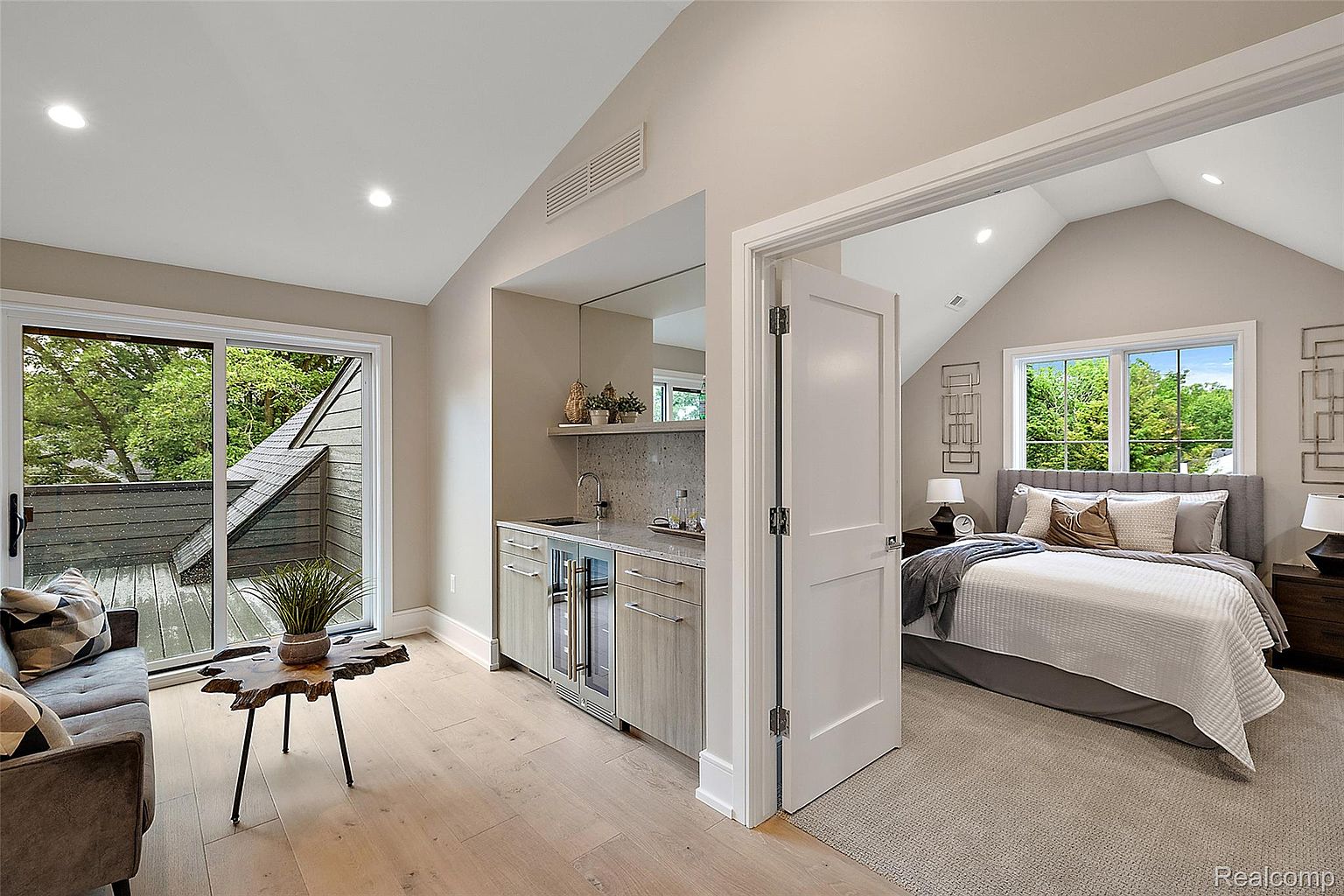 This image showcases a primary bedroom suite featuring a wet bar area adjacent to a bedroom with a vaulted ceiling. The bedroom includes a large window providing natural light, a bed with neutral bedding, and stylish wall decor. The wet bar area adds a touch of luxury and convenience to the suite.