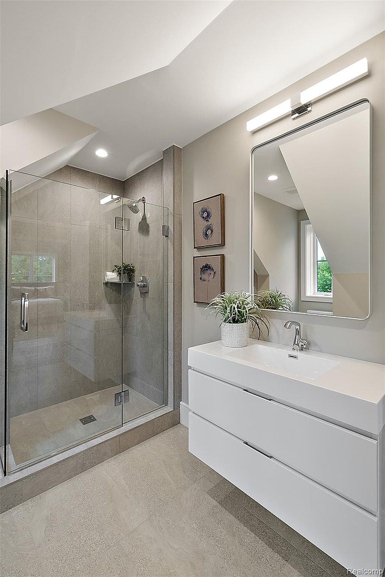 This is a well-lit primary bathroom featuring a glass-enclosed shower with gray tile and a modern white vanity with a sleek countertop and drawers. A large rectangular mirror hangs above the vanity, reflecting the room's clean lines and neutral color palette. The overall impression is one of contemporary elegance and functionality.