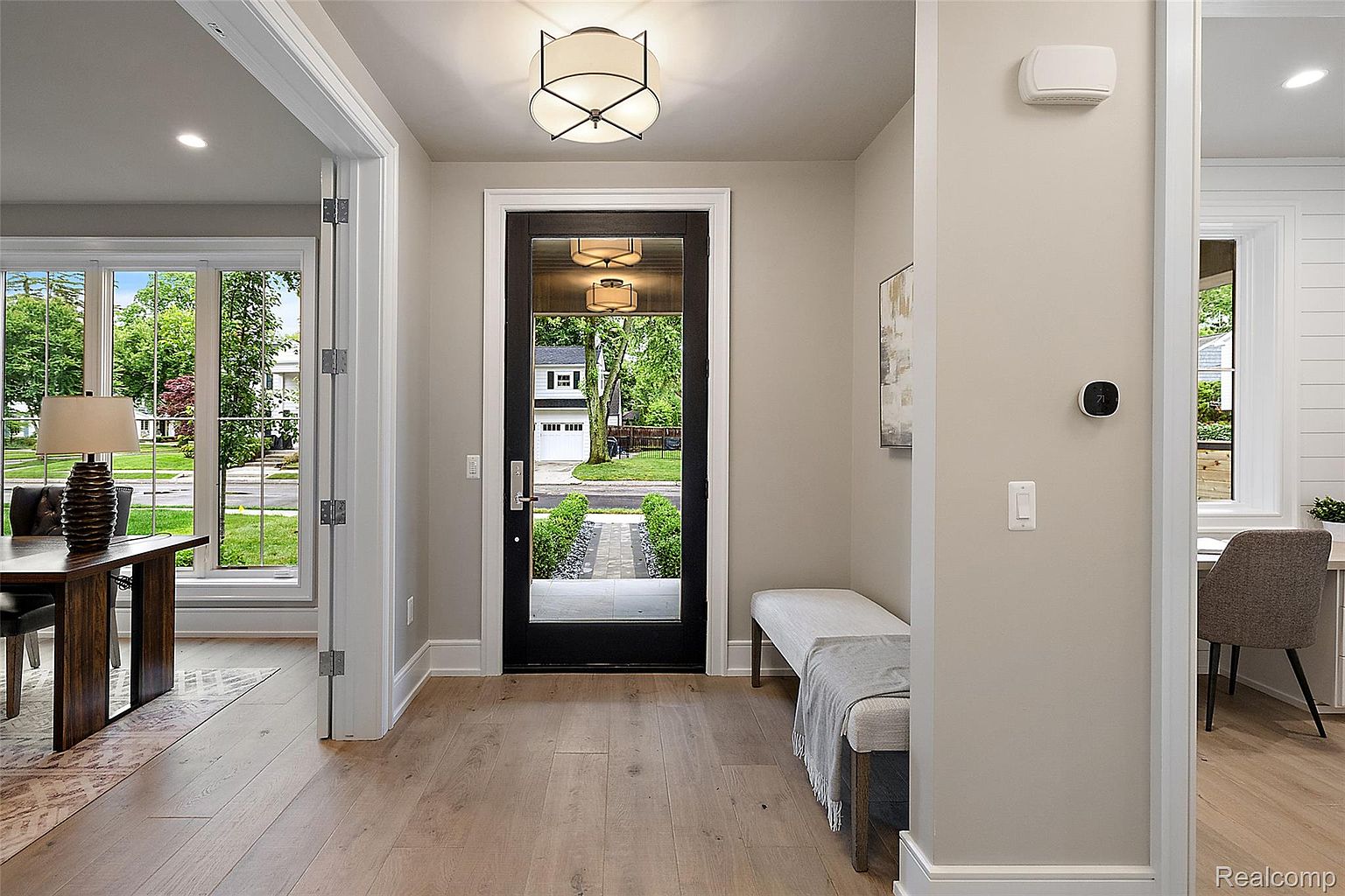 This interior shot showcases a modern hallway with light hardwood flooring and neutral-toned walls. A black-framed glass door provides a view to the exterior, while a bench with a throw blanket offers a seating area. The space is well-lit with a contemporary ceiling fixture, creating a welcoming and stylish entryway.