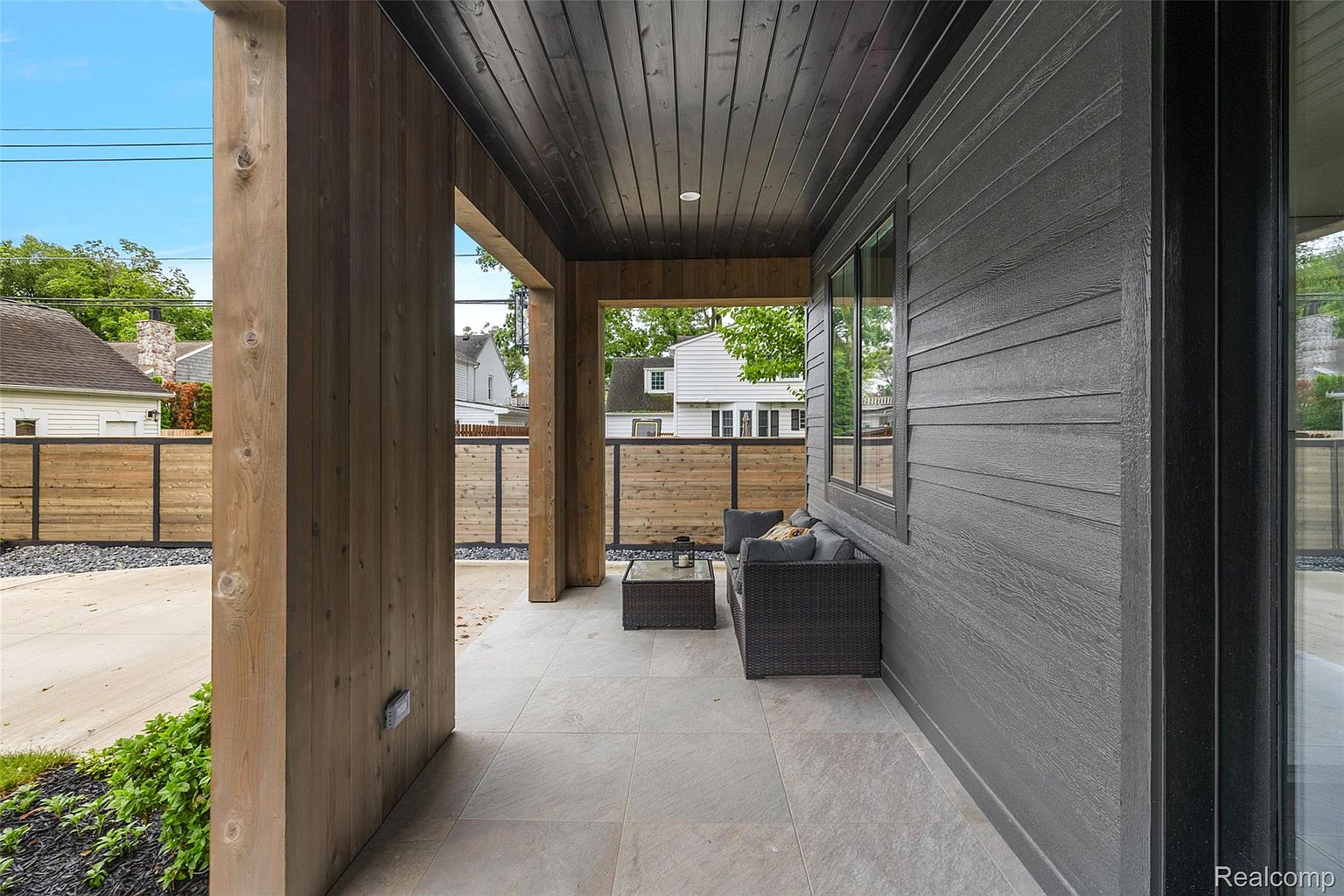 This image showcases a modern patio area with a sleek design. The patio features a gray tiled floor, a dark gray exterior wall with windows, and wooden support beams. A wicker sofa and glass-topped table provide a comfortable seating area, while a wooden fence offers privacy. The overall impression is one of contemporary elegance and outdoor relaxation.