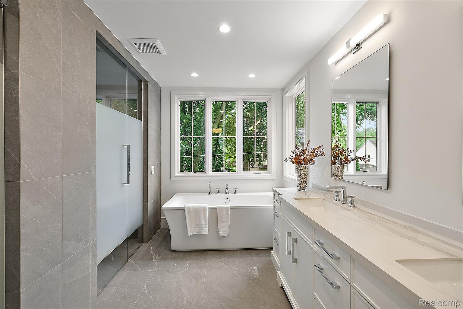 This is a bright and modern primary bathroom featuring a large soaking tub positioned beneath a window offering a view of lush greenery. The bathroom includes a spacious vanity with a white countertop, complemented by a large mirror and contemporary lighting fixtures. A glass-enclosed shower adds to the luxurious feel of the space, which is finished with elegant gray tile flooring.