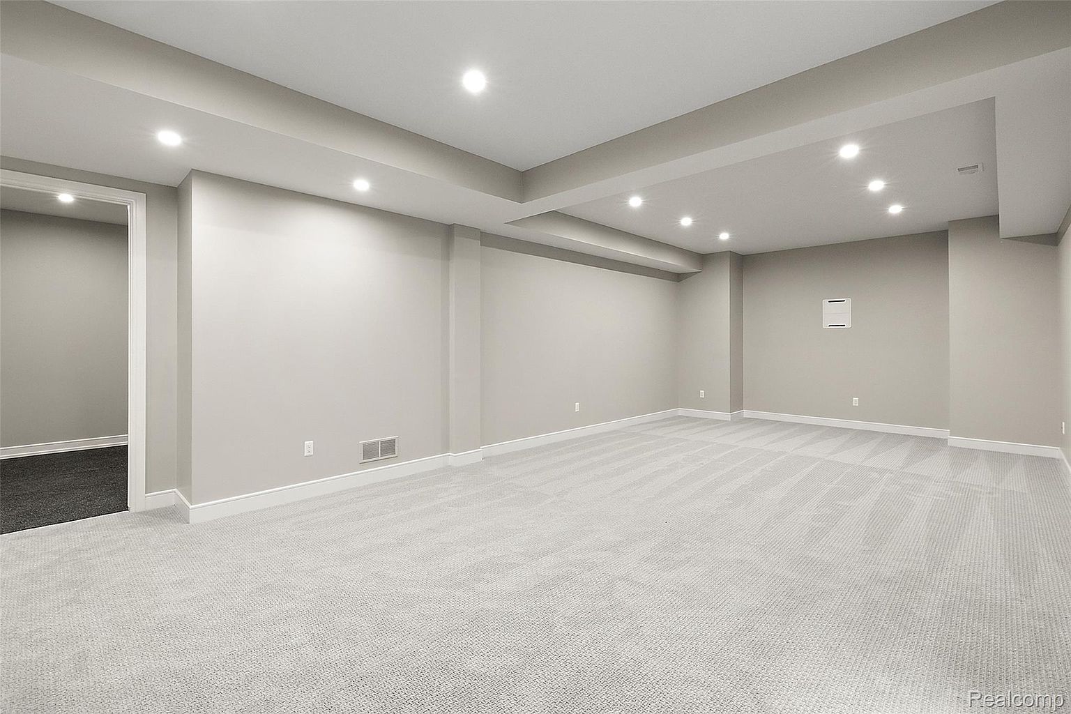 This is a spacious, finished basement with neutral-toned walls and light gray carpeting. Recessed lighting provides ample illumination, and the room features clean lines and a minimalist design. A doorway leads to another area, and the overall impression is one of a clean and versatile space.