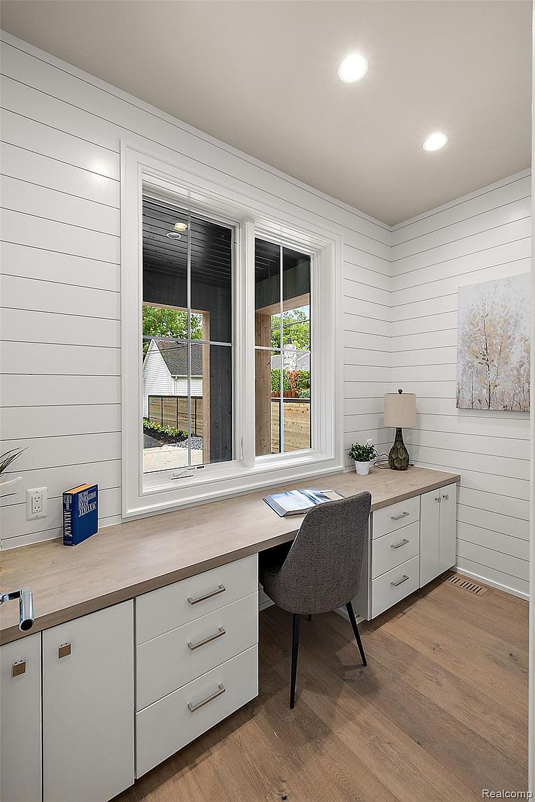 This is an interior shot of a home office or study. The room features white shiplap walls, a built-in desk with light wood countertop, and white cabinets with drawers. A large window provides natural light and a view of the outdoors, while a gray chair sits at the desk. A lamp and artwork add to the room's decor.
