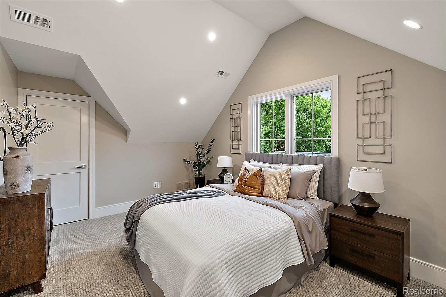 This is a cozy primary bedroom featuring a bed with a gray upholstered headboard, neutral-toned bedding, and decorative pillows. The room has an angled ceiling, a window providing natural light, and a dark wood nightstand with a lamp. The overall style is comfortable and inviting, perfect for relaxation.