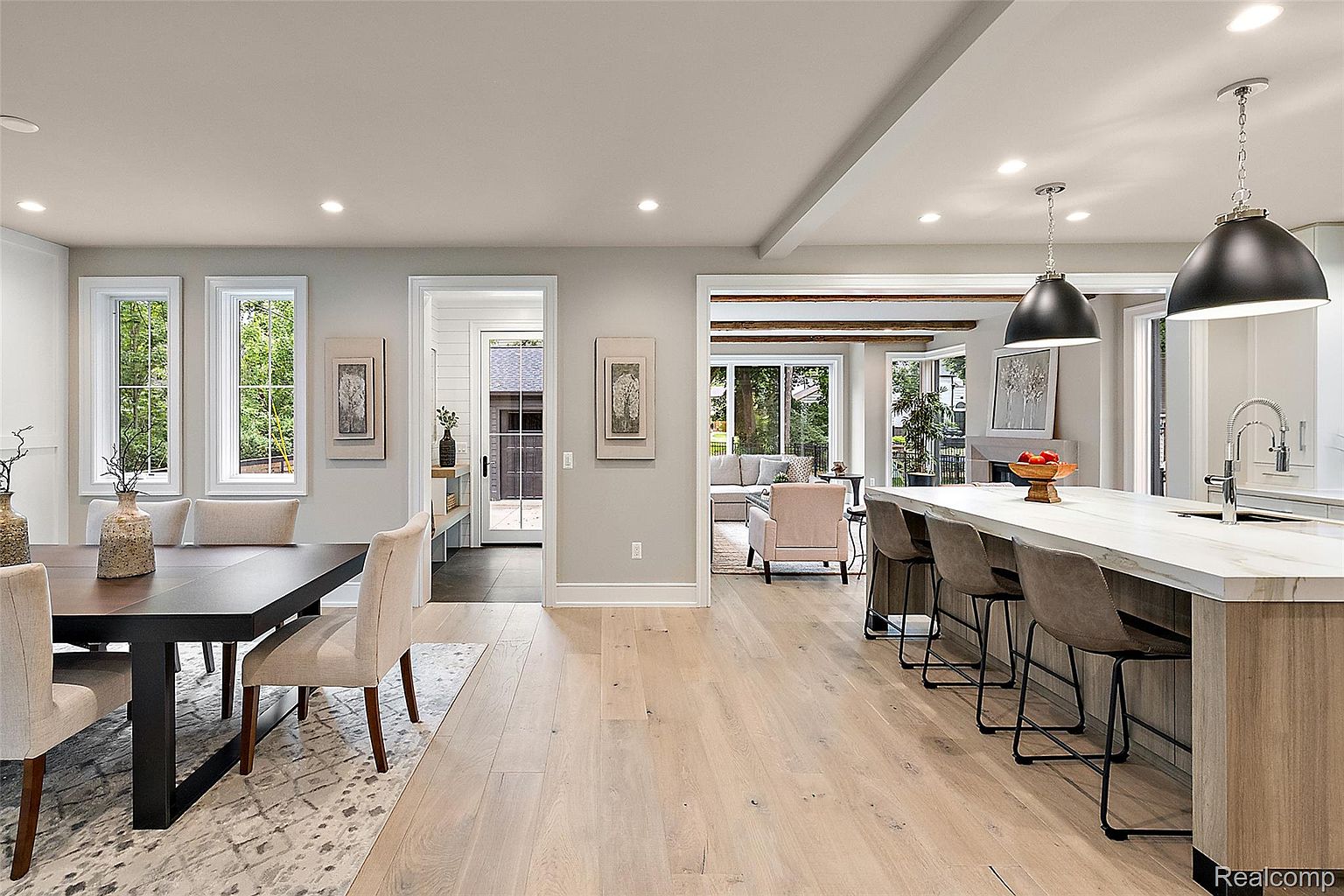 This interior shot showcases a modern kitchen and dining area with an open floor plan. The kitchen features a large island with bar seating and pendant lighting, while the dining area includes a dark wood table with upholstered chairs and a patterned rug. Natural light floods the space through large windows, creating a bright and inviting atmosphere.