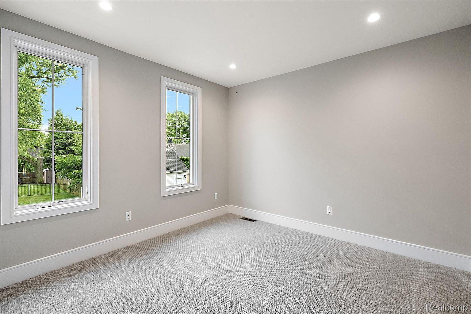 This is a well-lit, empty bedroom with neutral gray walls and light gray carpet. Two windows provide natural light and views of the outside greenery. The room is clean and ready for personalization, offering a blank canvas for potential buyers.