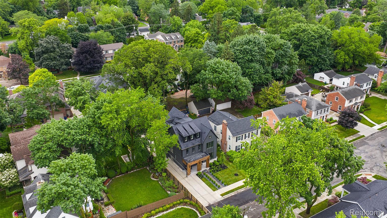 This aerial view showcases a modern, dark-colored home nestled among lush greenery in a suburban neighborhood. The property features a well-manicured lawn, a wooden fence, and a paved driveway leading to a two-car garage. The surrounding mature trees provide privacy and a sense of established community.