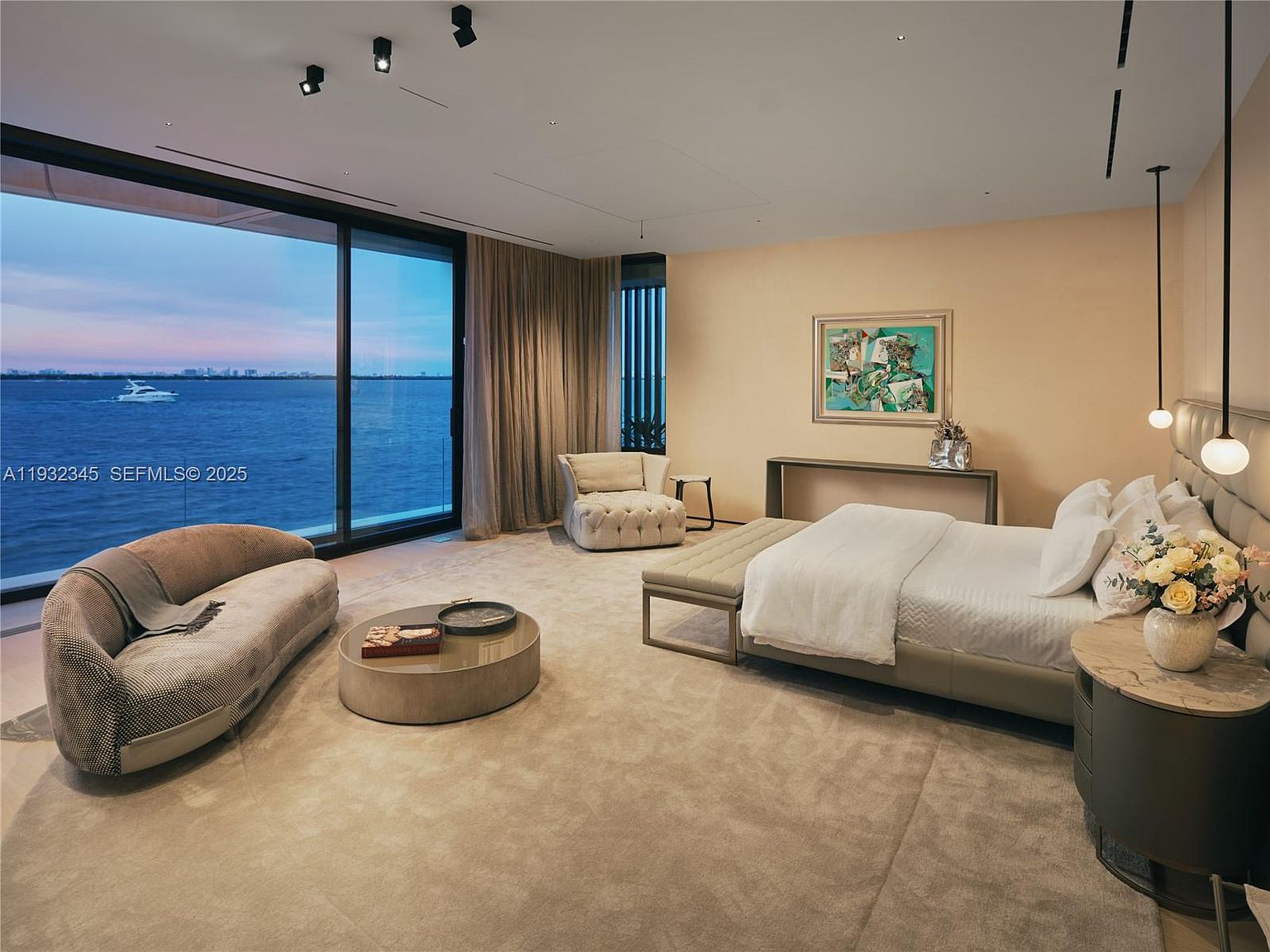 This is a luxurious primary bedroom featuring a large window with a waterfront view, a comfortable seating area with a curved sofa and round coffee table, and a king-size bed with a padded headboard. The room is decorated in neutral tones with a beige carpet and walls, creating a serene and inviting atmosphere. The perspective is from the doorway, showcasing the entire room.