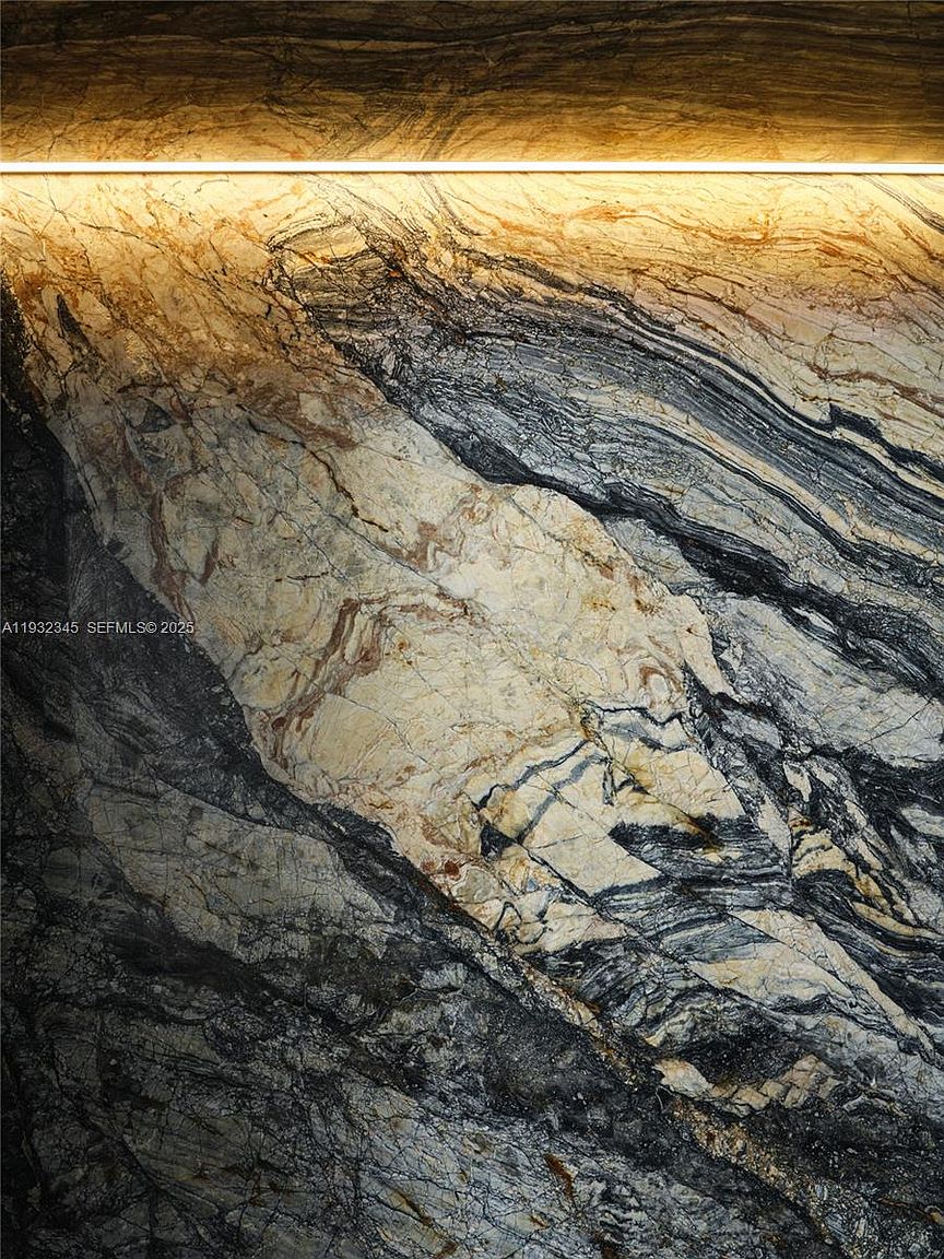 The image showcases a striking architectural detail, likely a wall or countertop made of intricately patterned marble. The stone features a blend of light beige and dark gray tones, creating a dramatic and luxurious effect. A warm light source illuminates the top portion, highlighting the texture and depth of the marble, adding to its visual appeal.