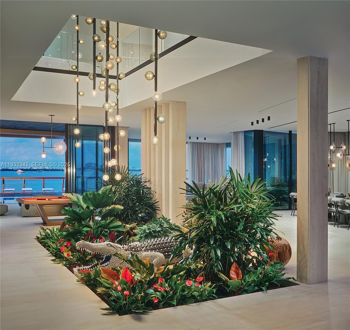 This is a luxurious living room featuring a unique indoor garden with an alligator sculpture as a focal point. The room boasts high ceilings with modern pendant lighting, large windows offering ocean views, and a neutral color palette. The overall impression is one of sophisticated elegance and contemporary design.