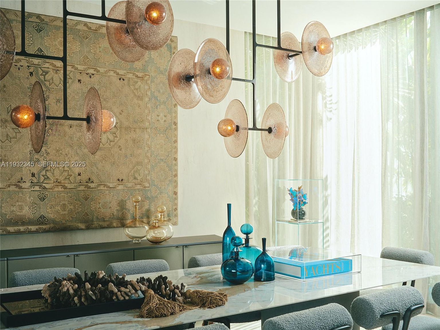 This interior shot showcases a sophisticated dining room featuring a long marble table adorned with decorative glass bottles and a coral centerpiece. Above the table, a modern chandelier with disc-shaped shades provides ambient lighting. A large patterned tapestry hangs on the wall, adding texture and visual interest to the space.