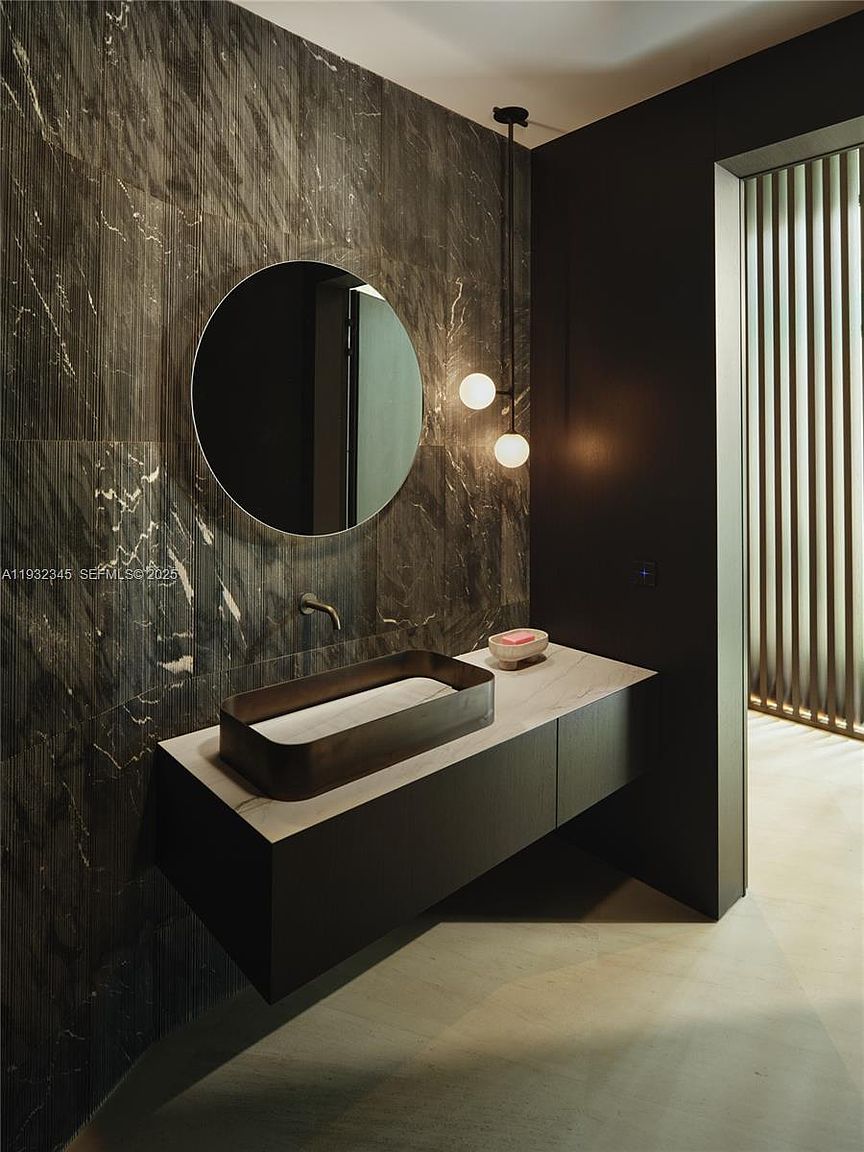 This is a modern bathroom featuring a dark, moody aesthetic. The walls are covered in a textured, dark marble-like material, complemented by a round mirror and a unique, rectangular vessel sink. A floating vanity with a light countertop adds to the contemporary feel, while pendant lighting provides a soft glow, creating a luxurious and sophisticated space.