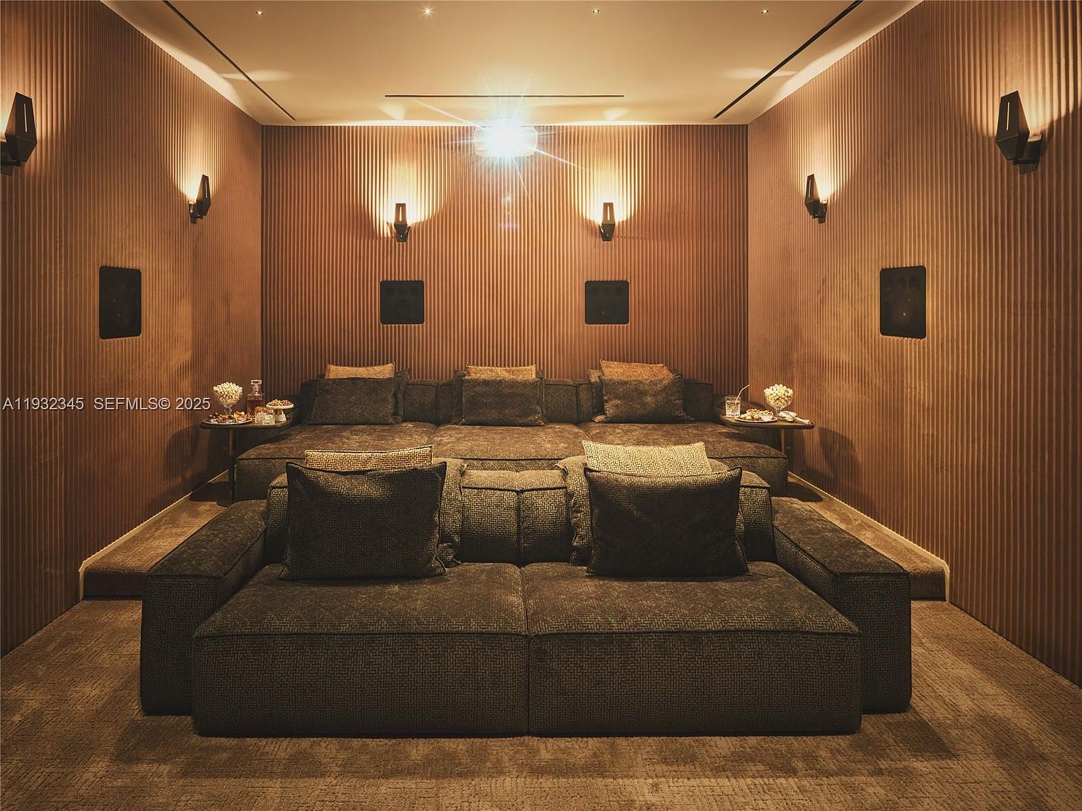 This is an interior shot of a home theater room. The room features tiered seating with comfortable, dark-colored couches and throw pillows. The walls are paneled with vertical wood slats and sconce lighting, creating a warm and inviting atmosphere. The perspective is from the front row, looking towards the screen, emphasizing the immersive experience.