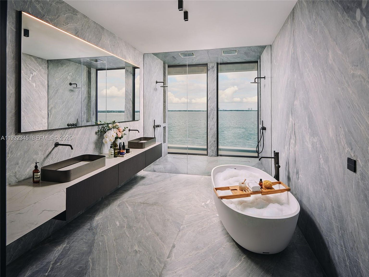 This is a luxurious primary bathroom featuring a modern design with gray marble walls and flooring. The room includes a double vanity with sleek, dark-toned cabinets and rectangular sinks, a large mirror with integrated lighting, and a freestanding bathtub filled with bubbles, positioned near a window offering a scenic water view. The overall impression is one of spa-like tranquility and high-end elegance.