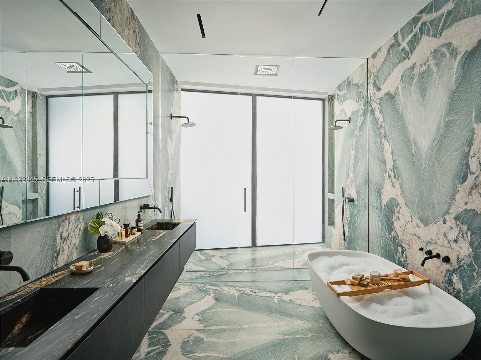 This is a luxurious primary bathroom featuring marble walls and flooring with striking veining. A freestanding soaking tub is positioned near a glass-enclosed shower area, and a long, dark vanity with dual sinks provides ample counter space. The overall impression is one of modern elegance and spa-like tranquility.