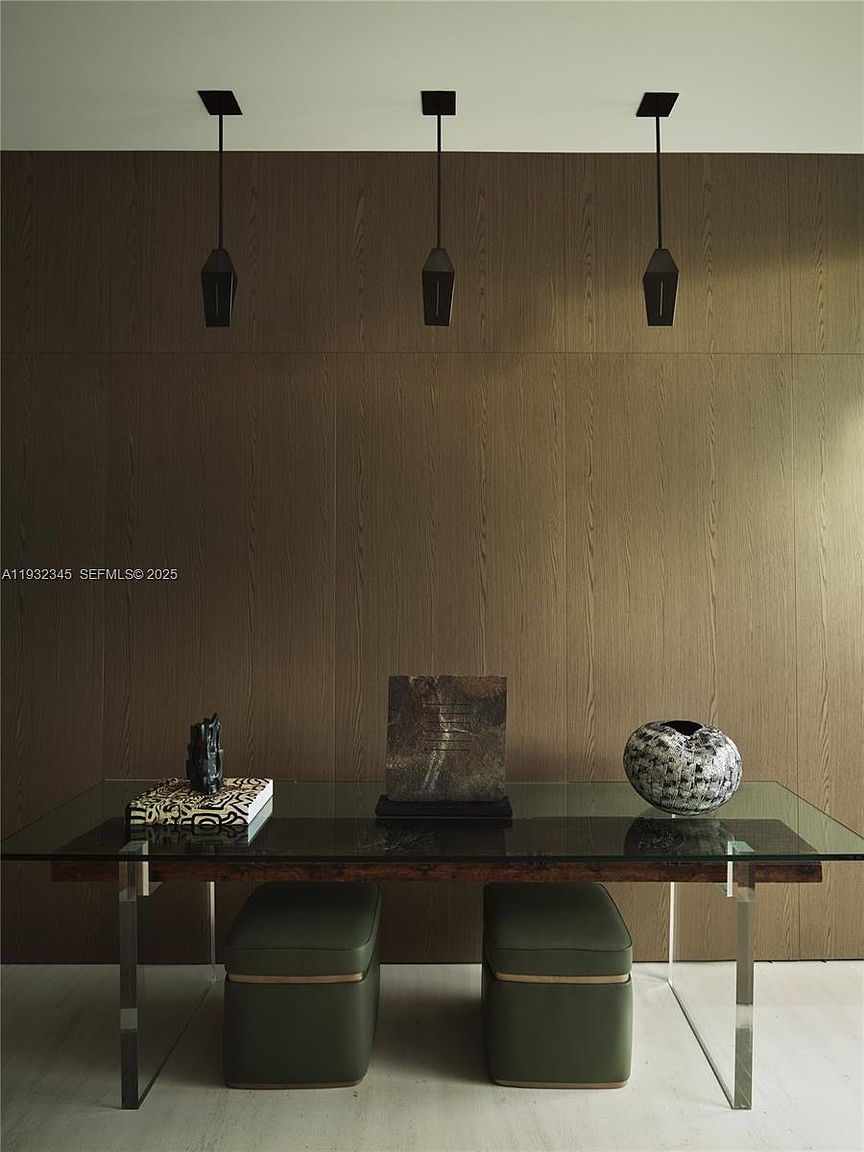 This interior shot showcases a modern dining room with a glass-topped table resting on a wooden base with metal legs. Two olive green ottomans are placed beneath the table. Above, three pendant lights hang against a wood-paneled wall, adding a touch of contemporary elegance.