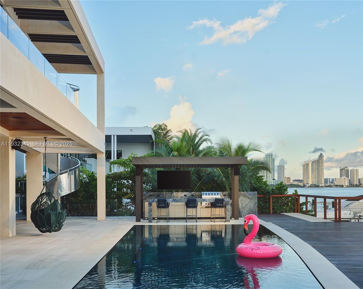 This image showcases a luxurious outdoor pool area with a modern design. The pool features a swim-up bar under a pergola with a television, and a pink flamingo float adds a playful touch. The area includes a wooden deck, a spiral staircase, and offers stunning views of the city skyline and waterfront, creating an inviting and upscale atmosphere.