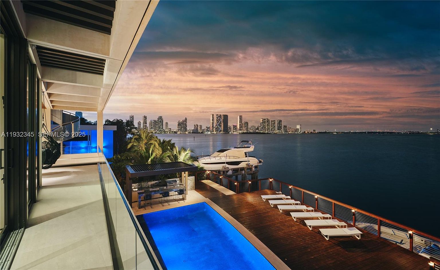 This image showcases a luxurious outdoor space featuring a stunning pool with vibrant blue water, a modern bar area, and a spacious wooden deck furnished with multiple lounge chairs. In the background, a sleek yacht is docked, and the city skyline is visible across the water, creating a picturesque and upscale ambiance perfect for relaxation and entertainment. The view is from a high angle, looking down on the pool and deck area.