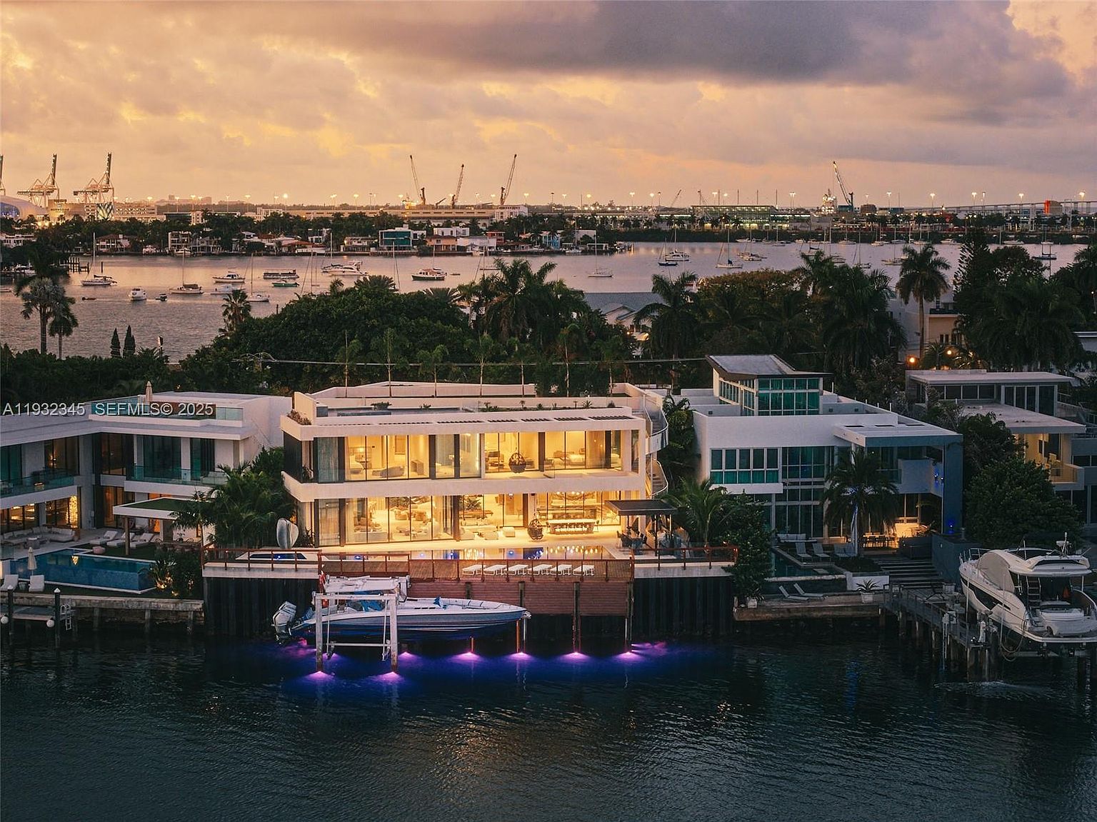 This aerial view showcases a luxurious waterfront property at dusk, featuring modern architecture with expansive glass windows and a private boat dock. The property includes multiple structures, a pool, and lush landscaping, all set against a backdrop of the city skyline and a tranquil bay dotted with boats, creating an impression of upscale coastal living.