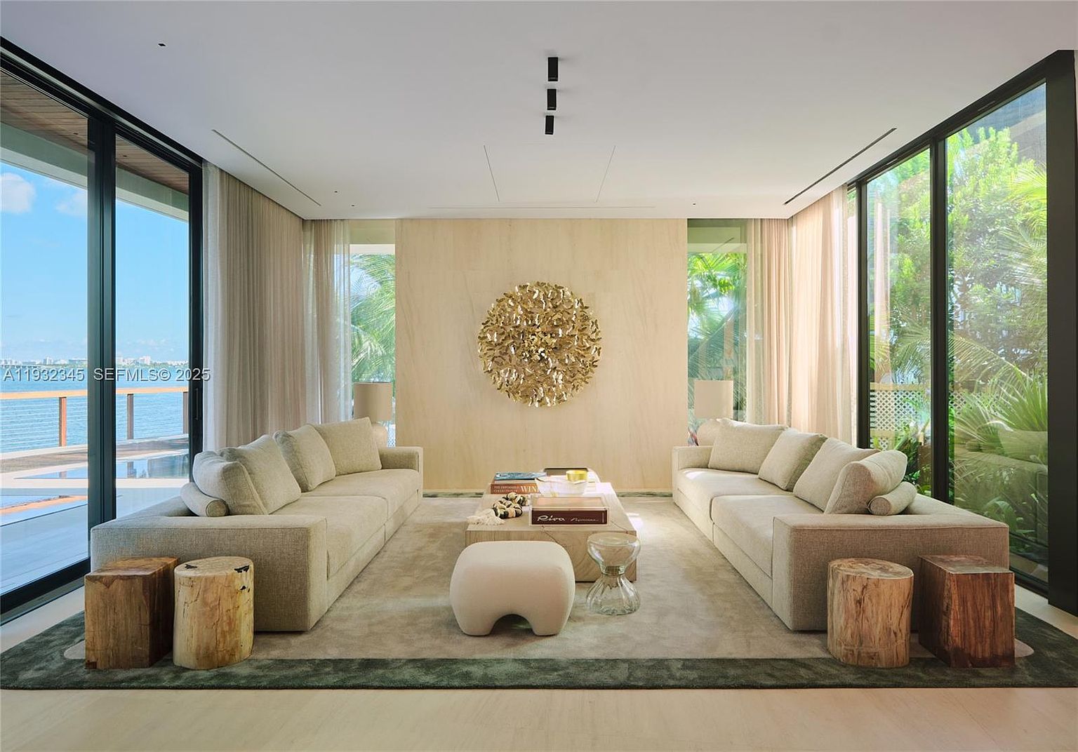 This is a spacious and elegant living room featuring two large sofas facing each other, separated by a coffee table and a unique ottoman. The room is decorated with a large, circular gold wall art piece and has floor-to-ceiling windows on either side, offering views of the outdoors. The color palette is neutral, creating a serene and sophisticated atmosphere.
