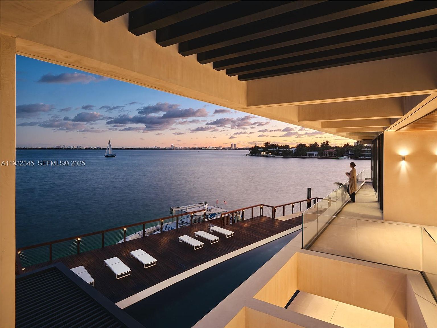 This image showcases a luxurious patio/deck/balcony area with modern design elements. The space features a wooden deck with lounge chairs, a sleek pool, and a glass railing offering stunning waterfront views. The architectural design includes a covered area with dark beams, creating a sophisticated and relaxing outdoor living space.