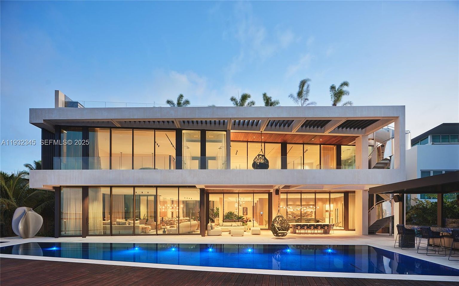 This is a rear exterior view of a modern, two-story luxury home featuring floor-to-ceiling windows and a flat roof with rooftop greenery. A large, rectangular pool with blue lighting is in the foreground, surrounded by a wooden deck. The home's design emphasizes indoor-outdoor living, with multiple seating areas visible through the windows and hanging chairs on the patio.