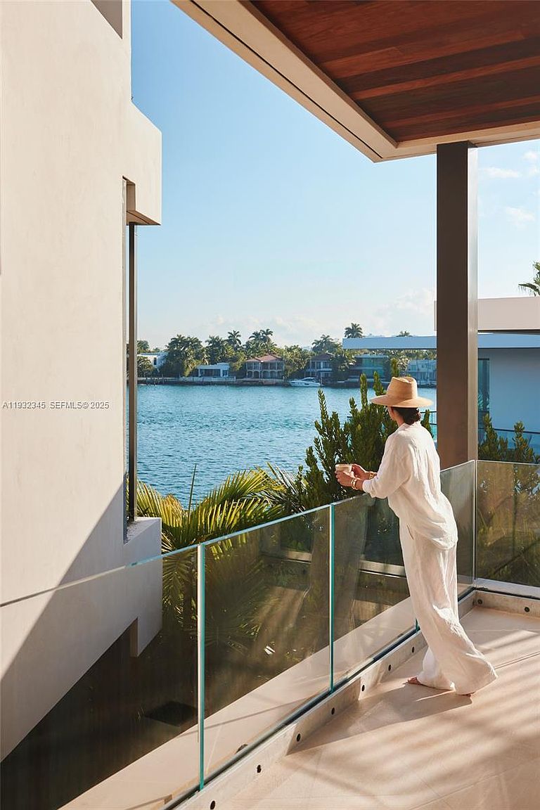 This image showcases a luxurious balcony with a stunning waterfront view. A woman in white attire and a hat is seen enjoying the scenery, emphasizing the relaxing atmosphere. The balcony features a modern glass railing and a sleek, tiled floor, enhancing the property's appeal for potential buyers seeking outdoor living spaces.
