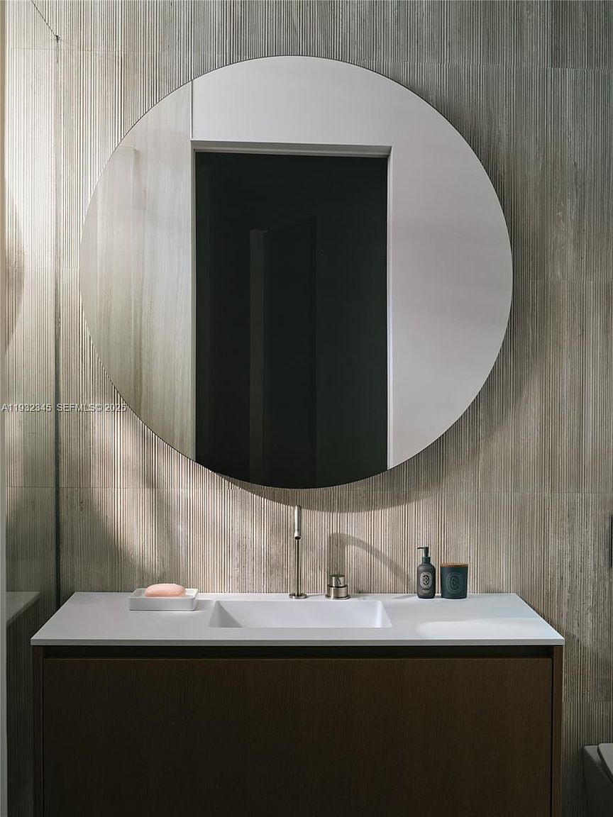 The image showcases a modern bathroom vanity with a round mirror. The vanity features a dark wood cabinet and a white countertop with an integrated sink. A sleek faucet and soap dispenser add to the minimalist aesthetic, while the textured wall behind the mirror provides visual interest.