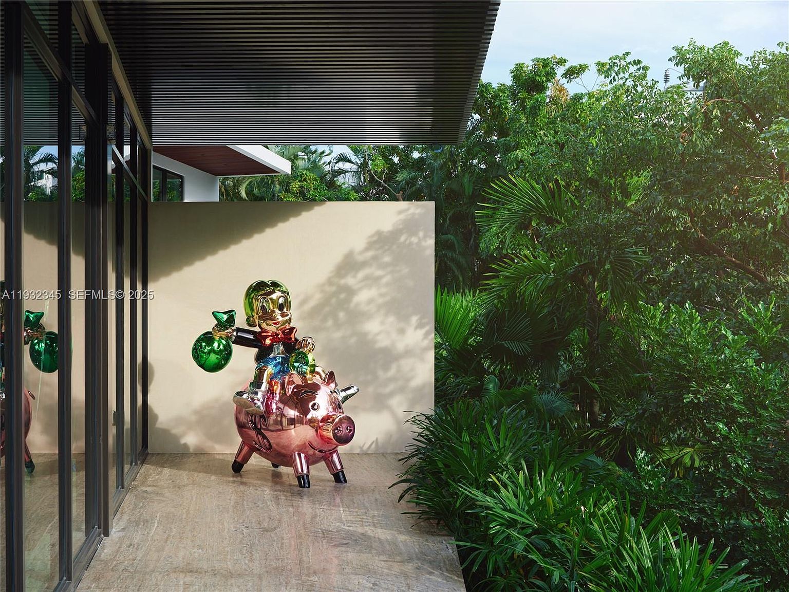 This image showcases a modern outdoor space featuring a whimsical sculpture of a boy riding a pig, adding a playful touch to the property. Lush greenery and mature trees create a private and serene garden setting. The architecture includes clean lines and a covered walkway, blending seamlessly with the natural surroundings.