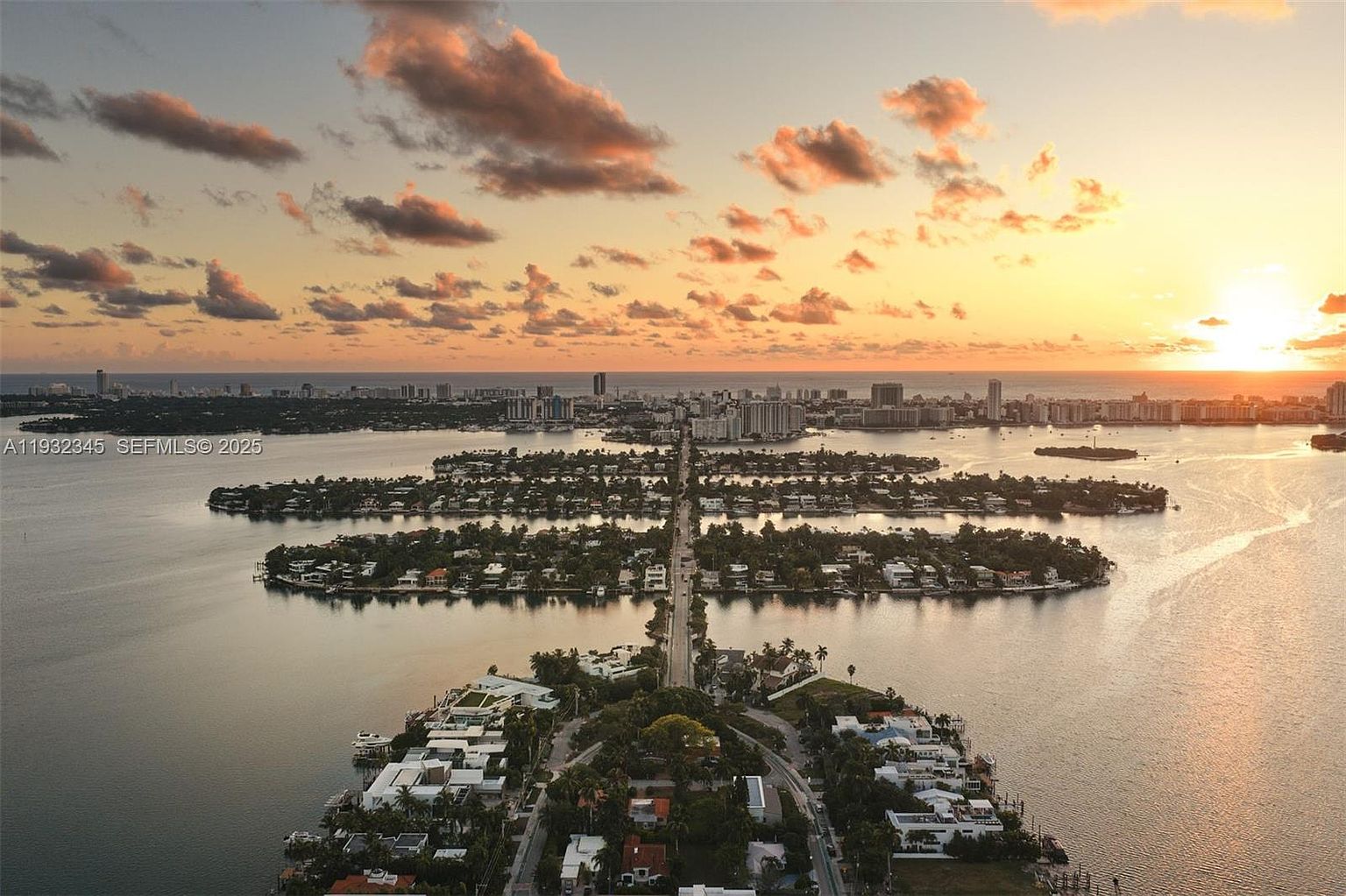 This aerial shot showcases a stunning waterfront property at sunset. The view captures a series of interconnected islands with luxury homes, a road bisecting the islands, and a cityscape in the distance under a sky filled with colorful clouds. The overall impression is one of exclusivity and high-end living.