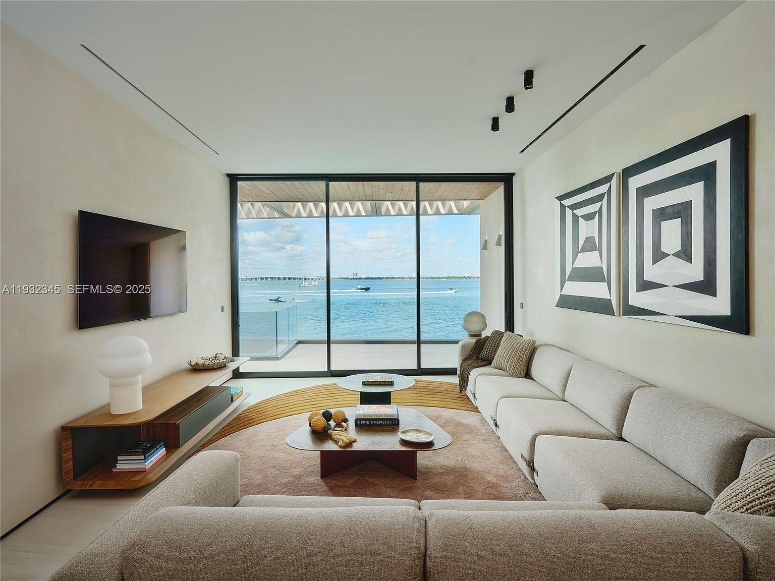 This is an interior shot of a modern living room with a neutral color palette. The room features a large sectional sofa, a low media console with a flat-screen TV, and floor-to-ceiling windows offering a waterfront view. Abstract art adorns the walls, and the space is well-lit, creating a serene and sophisticated atmosphere.