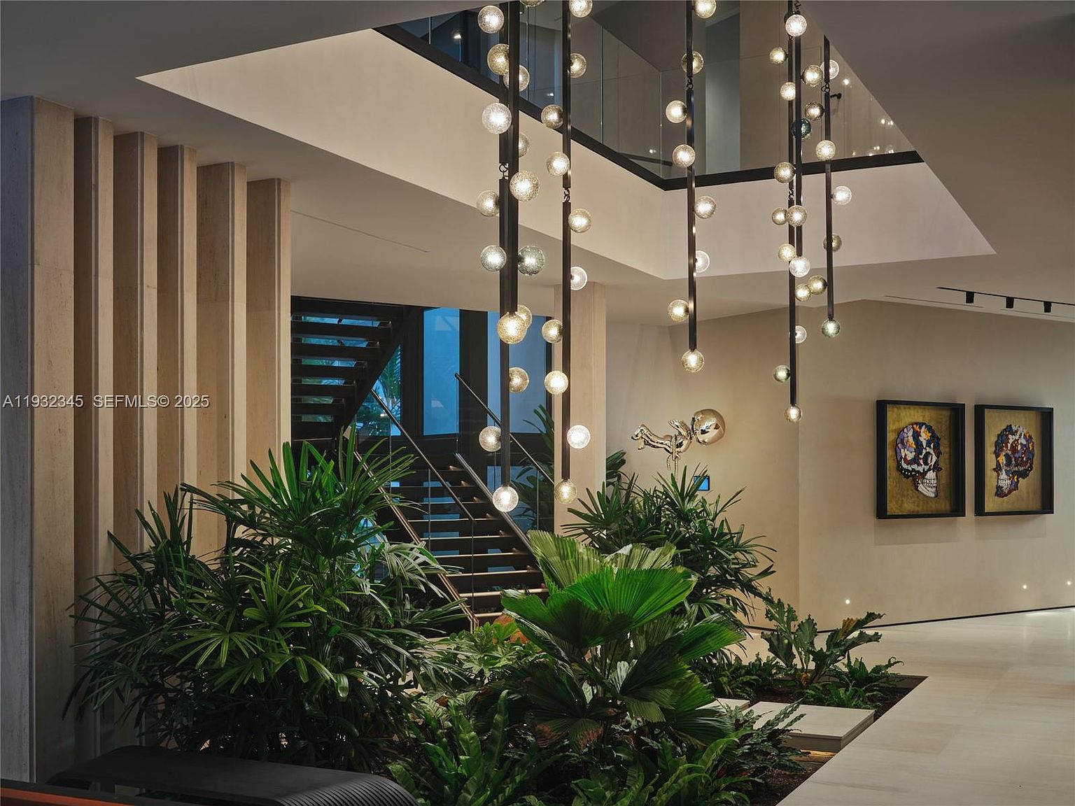 This interior shot showcases a modern hallway and staircase area, featuring a striking chandelier composed of multiple hanging lights. Lush greenery is incorporated into the design, adding a natural element to the contemporary space. The staircase leads to an upper level, visible through a large opening, and artwork adorns the walls, enhancing the sophisticated ambiance.