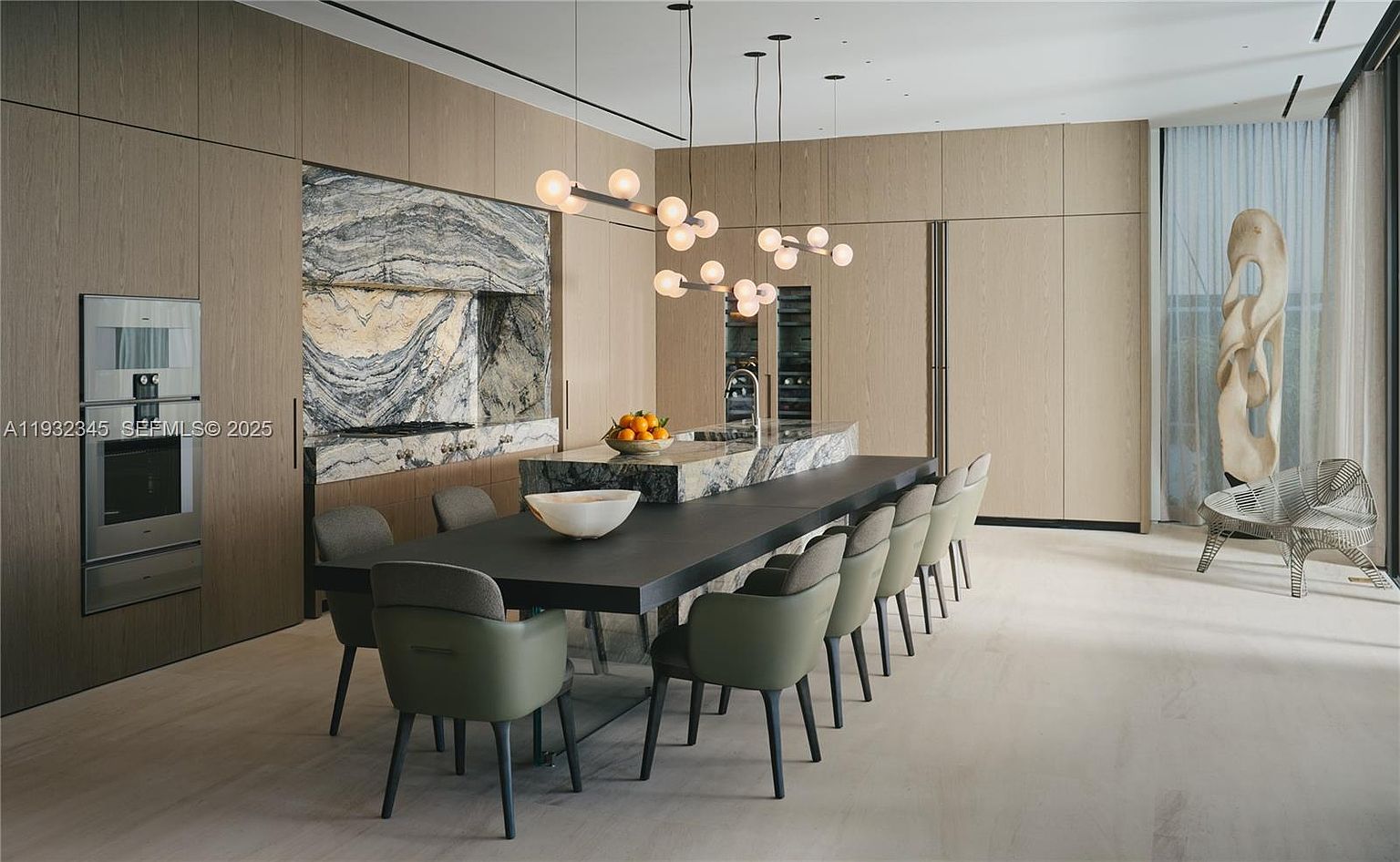 This interior shot showcases a modern dining room with a long, dark-colored table surrounded by green upholstered chairs. A unique lighting fixture hangs above the table, and a marble-clad wall feature adds a touch of luxury. The room also includes a sculpture and a wire chair near a window, creating an elegant and contemporary atmosphere.