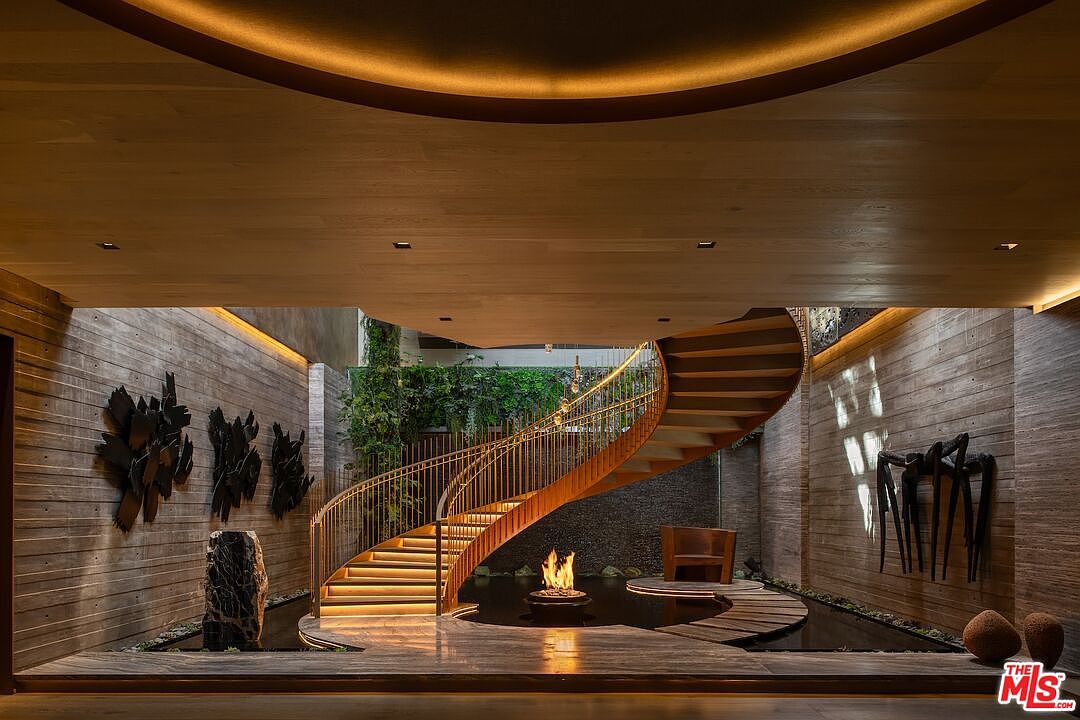 This interior shot showcases a unique hallway featuring a spiral staircase with warm lighting, complemented by a tranquil water feature and a fire pit. The walls are textured, adorned with artistic metal sculptures, and lush greenery adds a touch of nature. The perspective is from a low angle, emphasizing the height and grandeur of the space.