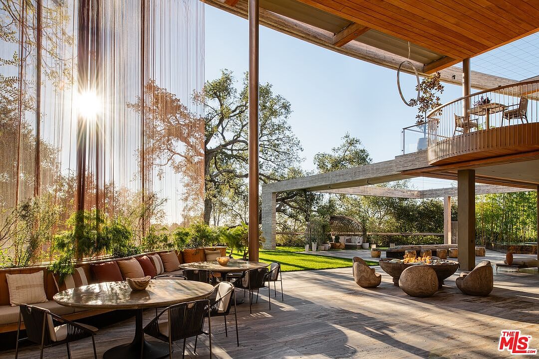 This image showcases a luxurious outdoor patio and deck area, featuring multiple seating arrangements including a built-in cushioned bench, round dining tables, and a fire pit surrounded by unique stone chairs. The space is partially covered by an architectural structure with wooden beams and a second-story balcony, blending seamlessly with the natural landscape and offering a sophisticated outdoor living experience. The sun shines through the trees and the vertical design element on the left, creating a warm and inviting atmosphere.